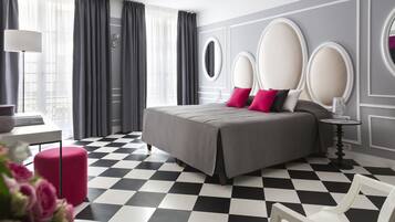 A modern hotel room with a checkered floor, a bed with a grey bedspread and pink pillows, a round mirror, and a small round table with a lamp.