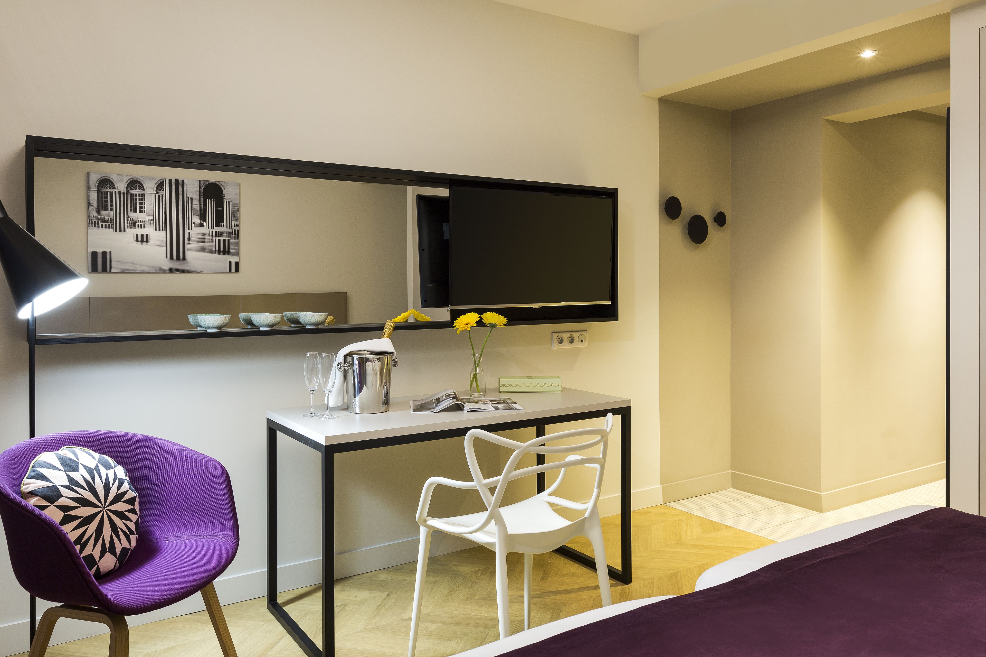 Une chambre d’hôtel moderne équipée d’un lit, d’un bureau avec un ordinateur, d’une chaise, d’une lampe et d’un téléviseur fixé au mur.