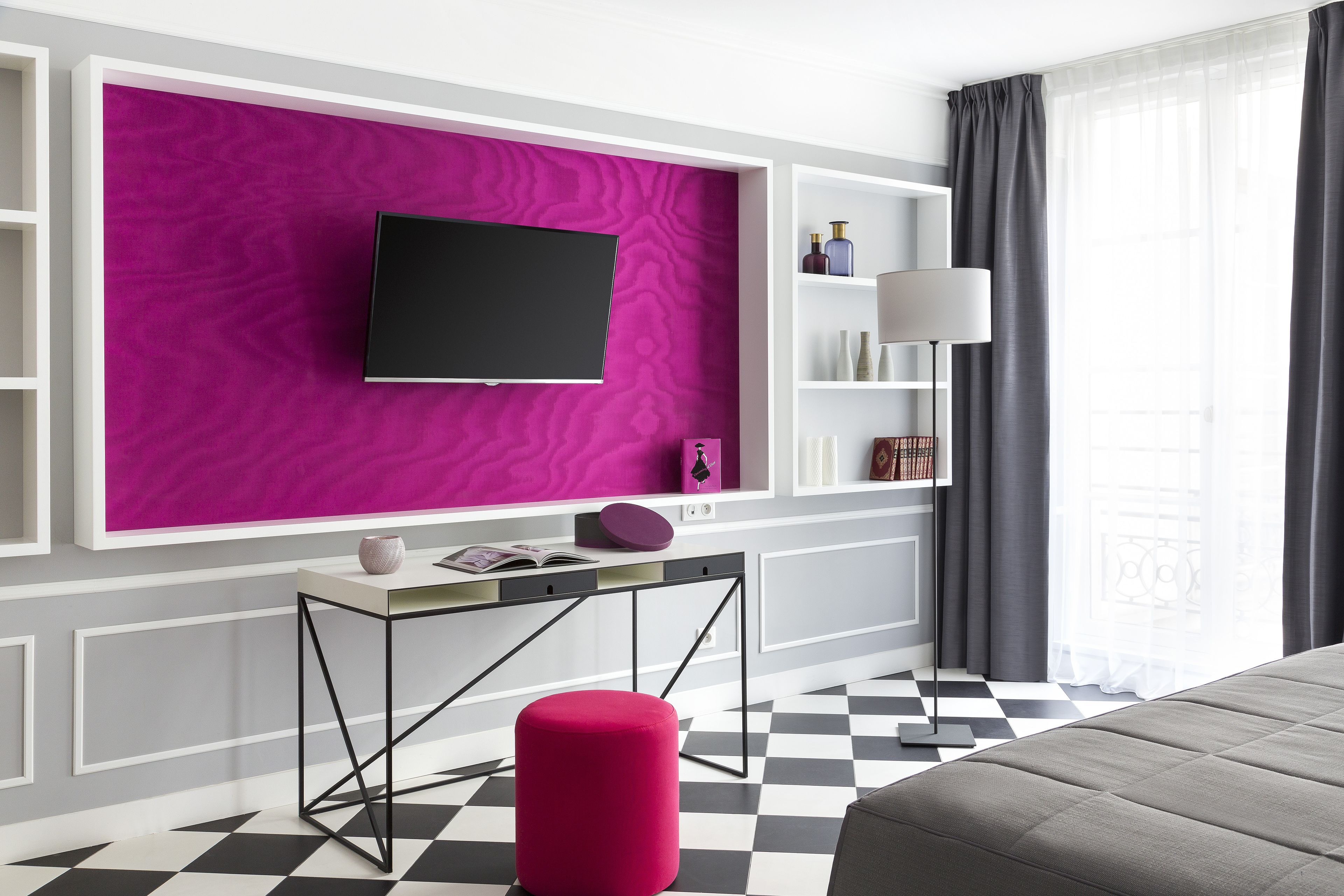 Une chambre d’hôtel moderne avec un sol à carreaux noir et blanc, un mur d’accent rose, un téléviseur à écran plat fixé au-dessus d’une console blanche et un canapé gris.
