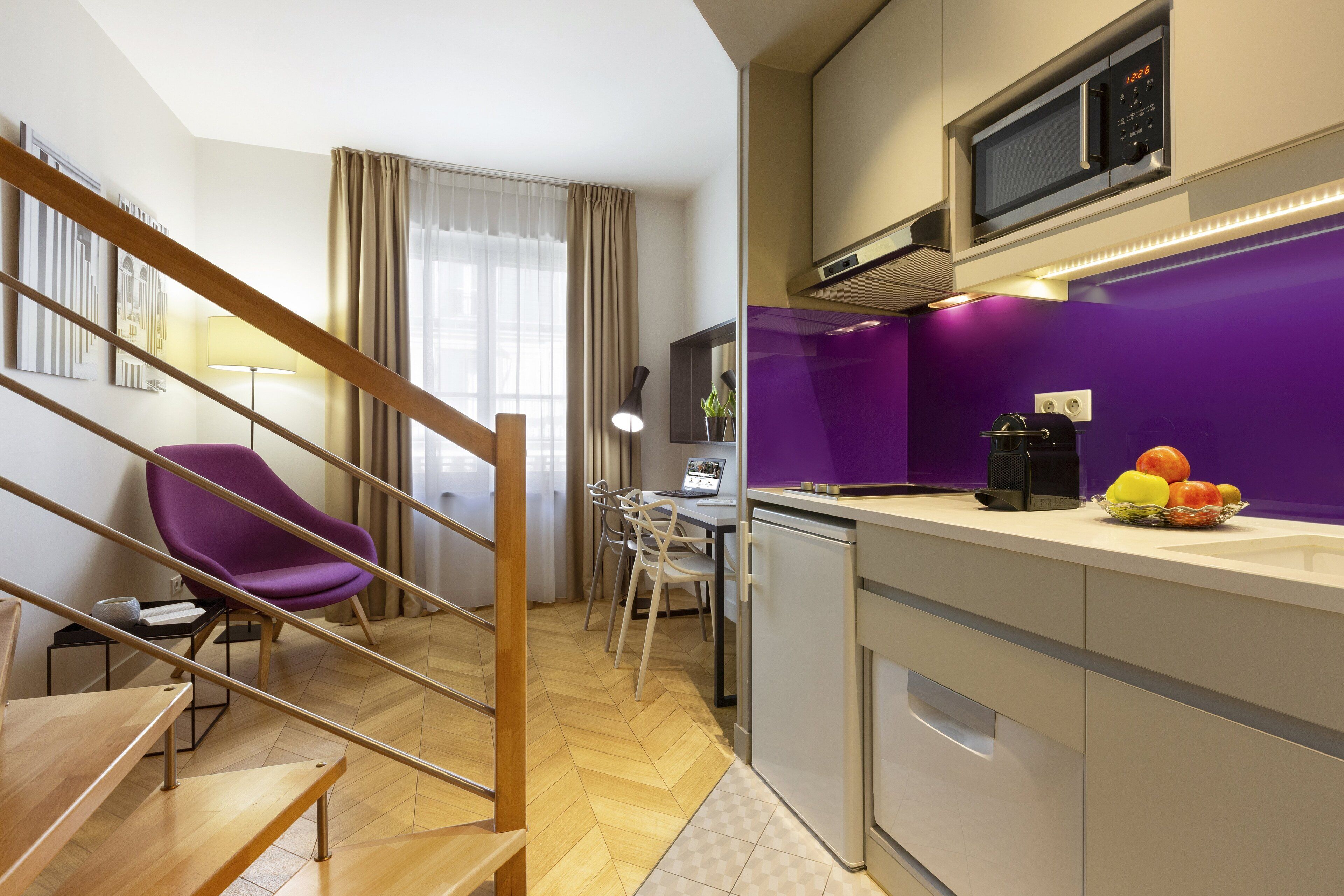 A modern kitchen with a microwave, a fruit bowl, and a dining area with a purple chair and a laptop on the table.