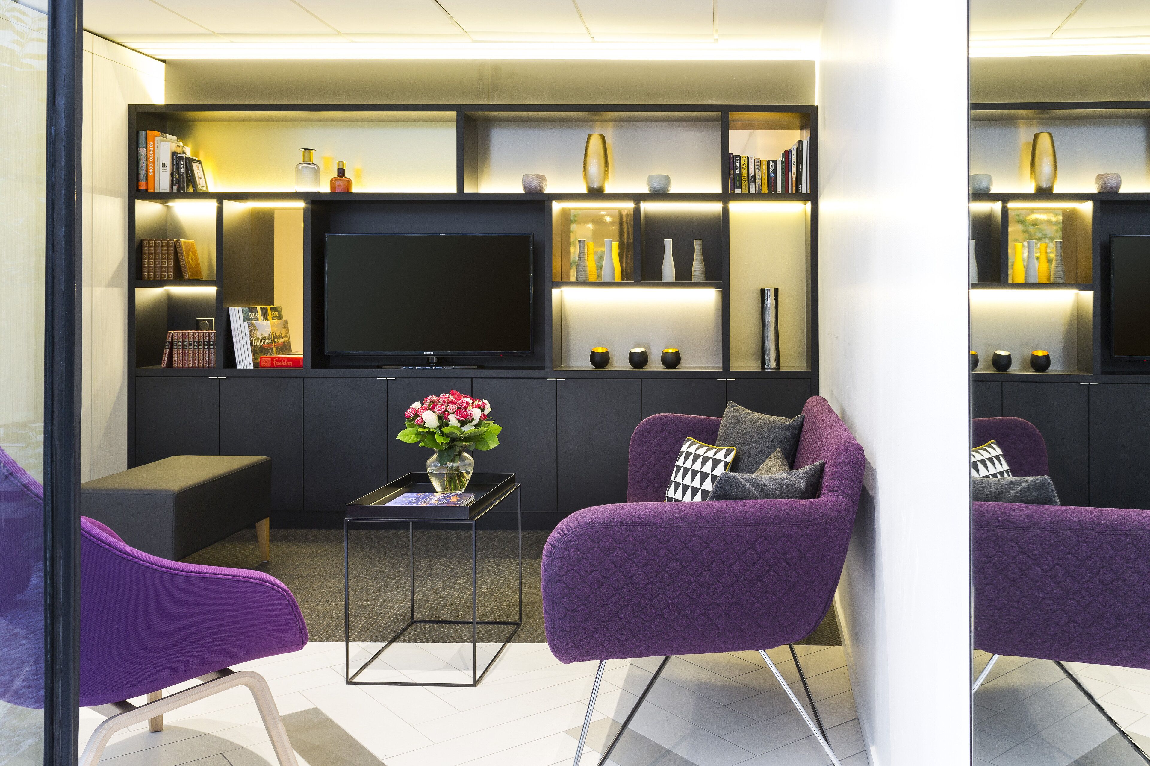 Un salon moderne doté d’un téléviseur à écran plat, de fauteuils violets, d’une table basse et d’une bibliothèque agrémentée d’objets décoratifs.
