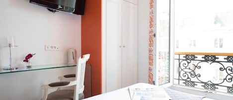 A hotel room with a bed, a TV mounted on the wall, a glass shelf with a vase, a white wardrobe, and a window with a balcony.