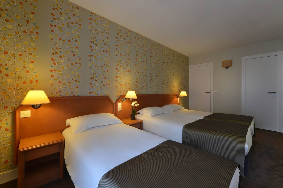A hotel room with two beds, a nightstand with a lamp, and a wall with a floral pattern.