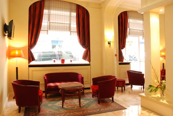 Un vestíbulo de hotel con mobiliario tapizado en rojo, una alfombra estampada y grandes ventanales con cortinas.
