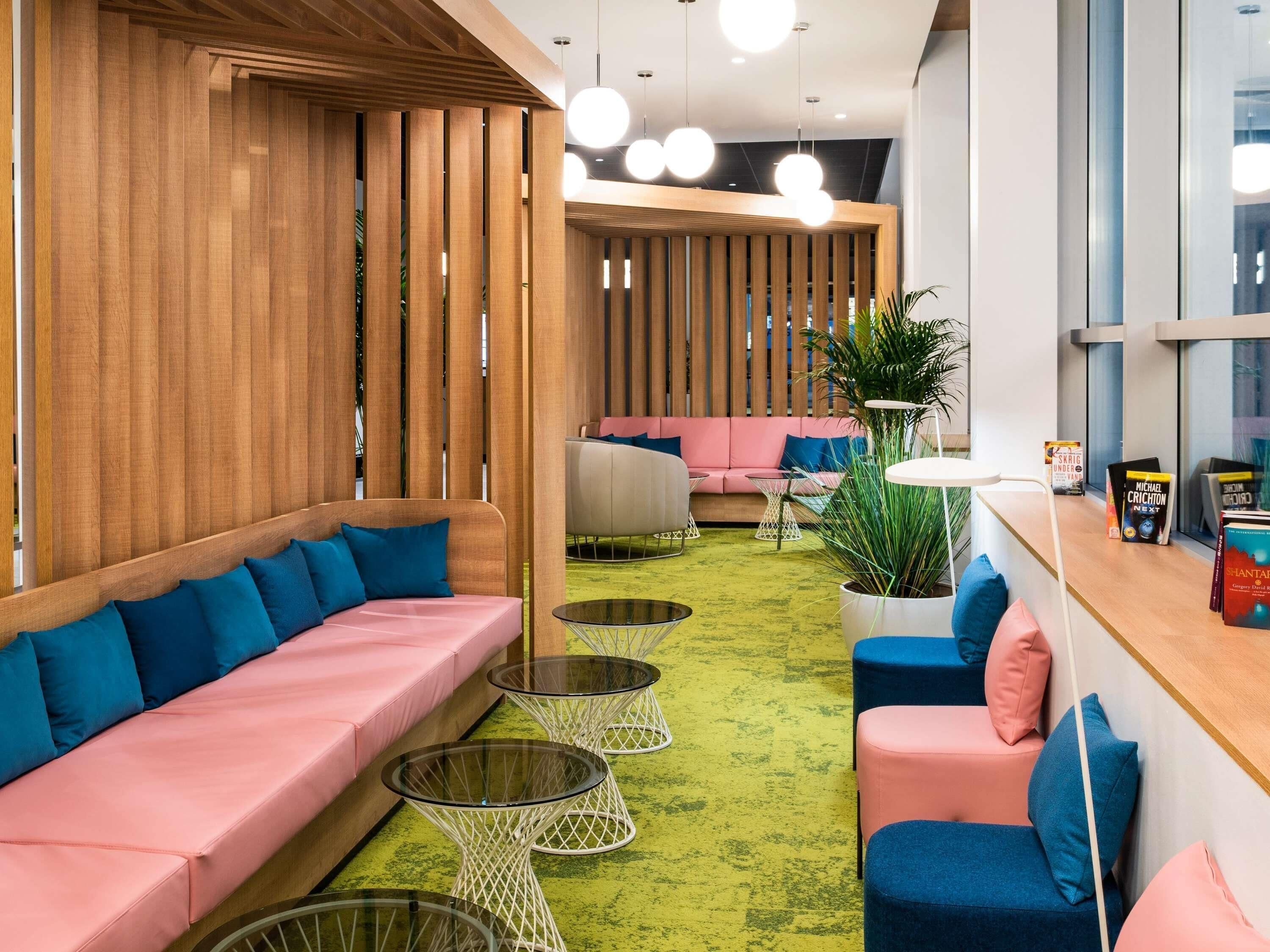 A modern lounge area with a green carpet, wooden partitions, and seating with blue and pink cushions.