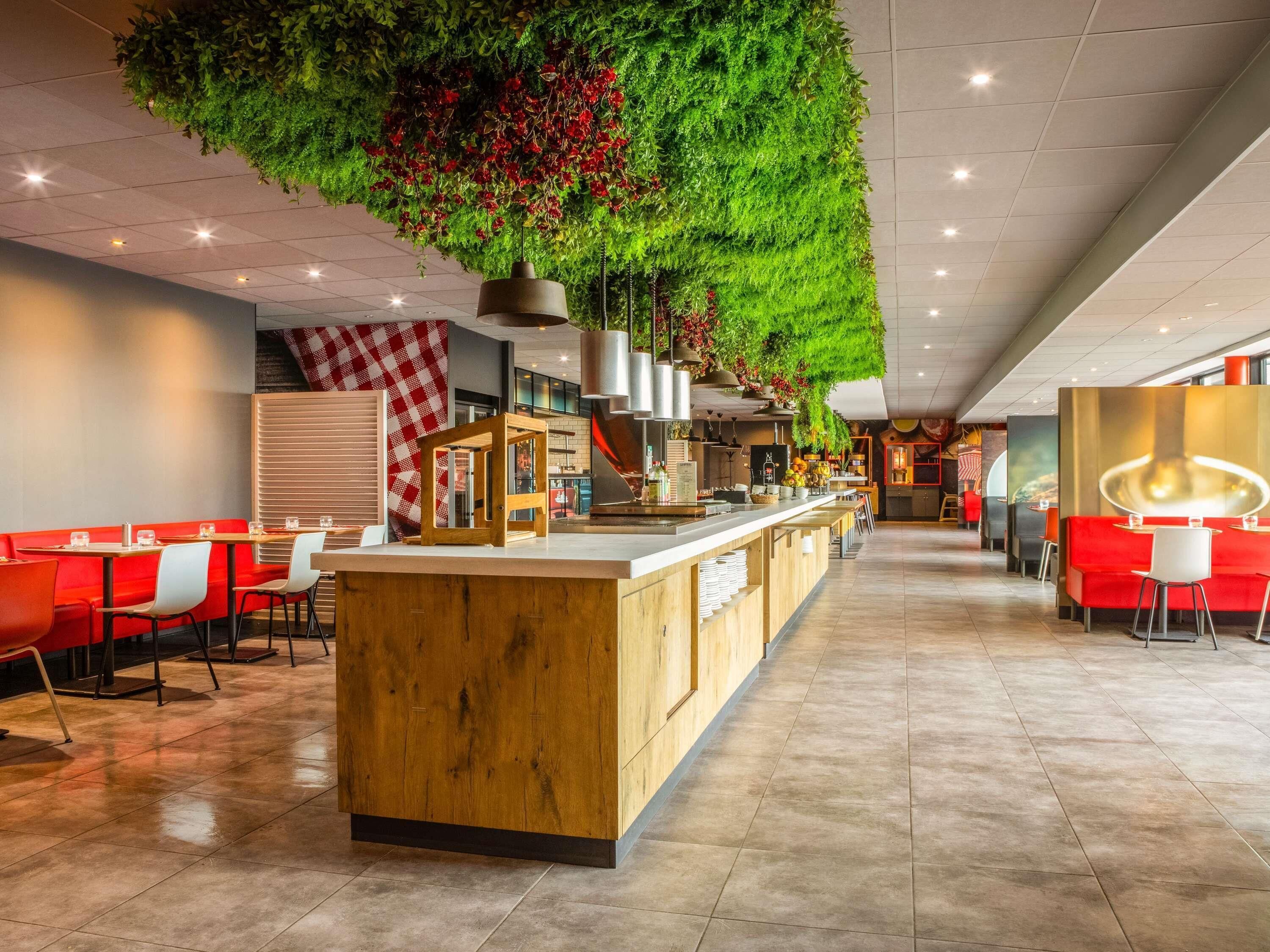 Un restaurante moderno con una barra central de madera, pared con cuadros rojos y blancos, y plantas colgantes verdes.