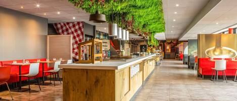 Un restaurante moderno con una barra central de madera, pared con cuadros rojos y blancos, y plantas colgantes verdes.