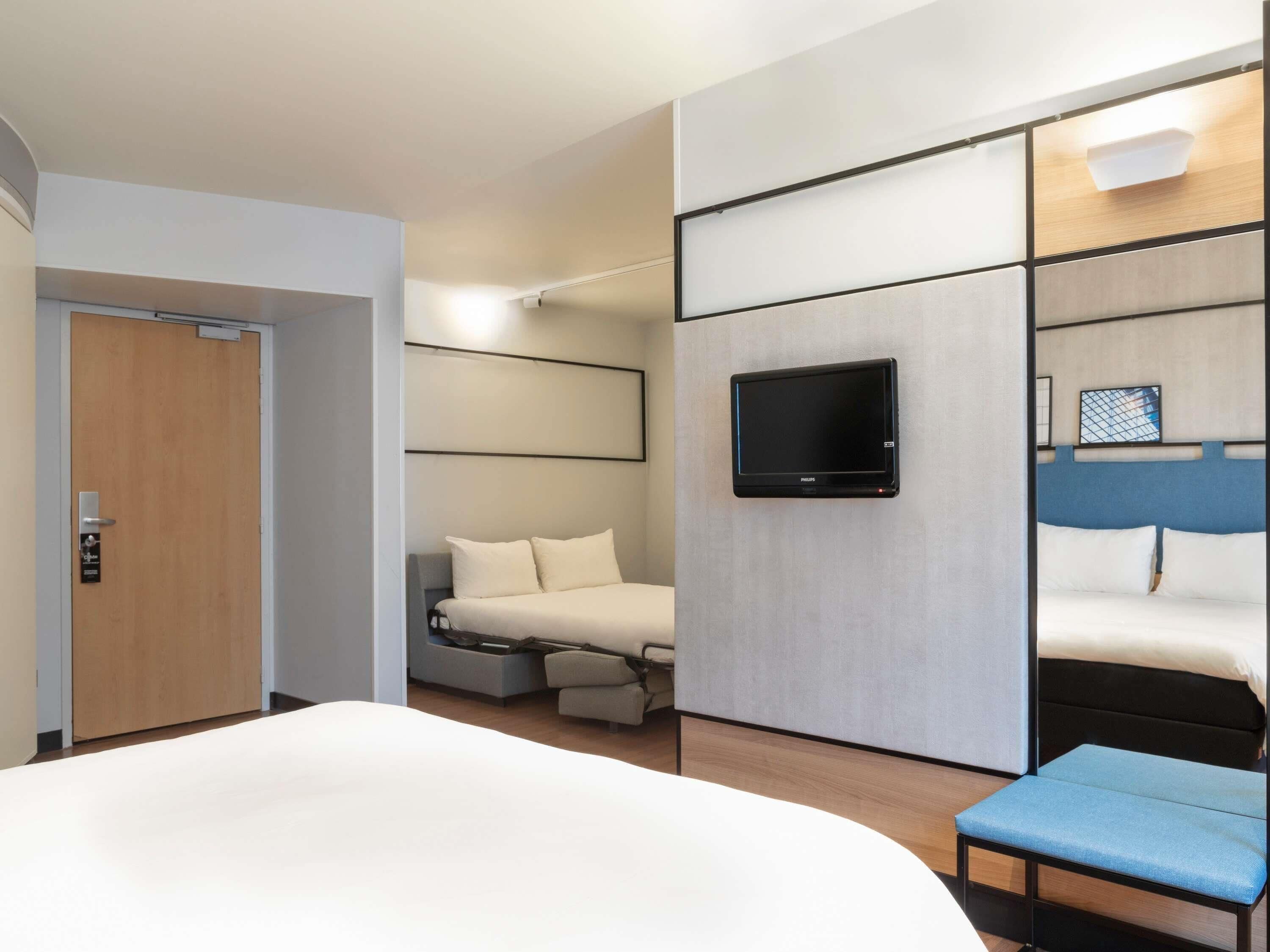 A modern hotel room with a large bed, a TV mounted on the wall, a small seating area, and a door.