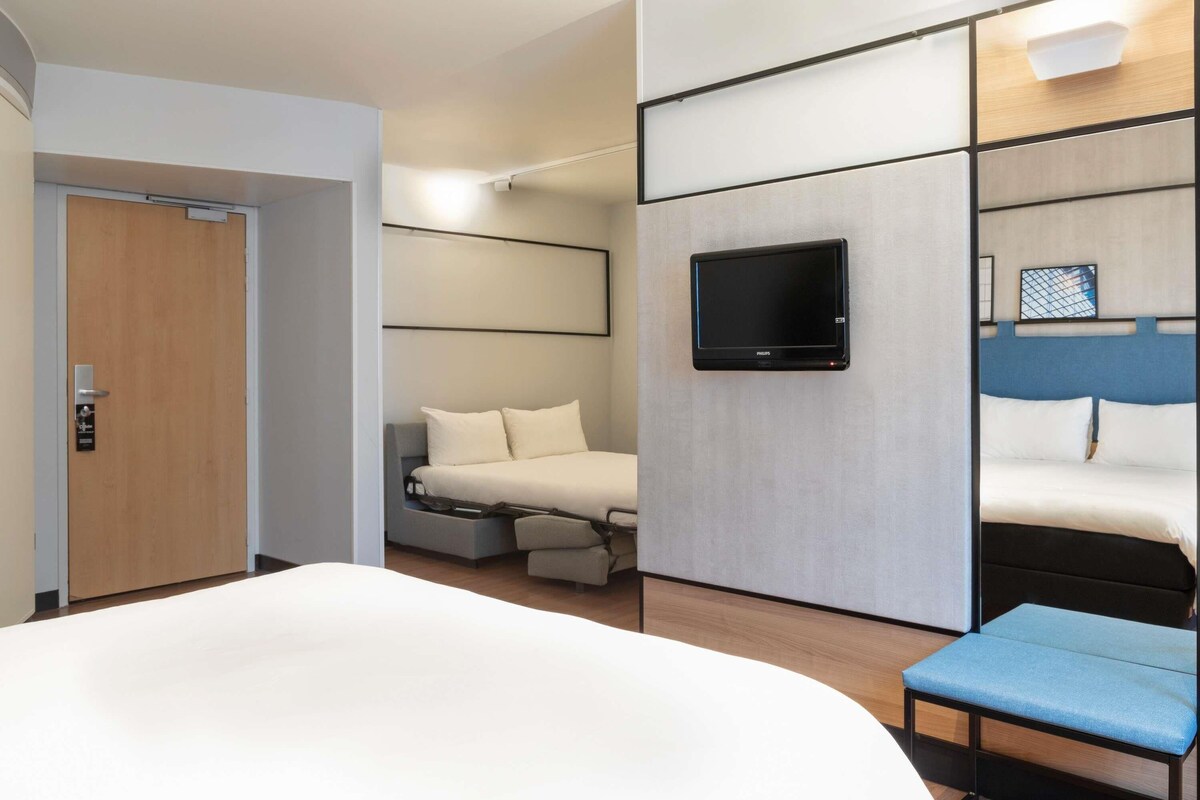 Una habitación de hotel moderna con una cama grande, un televisor montado en la pared, una pequeña zona de estar y una puerta.