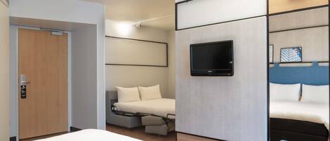 A modern hotel room with a large bed, a TV mounted on the wall, a small seating area, and a door.