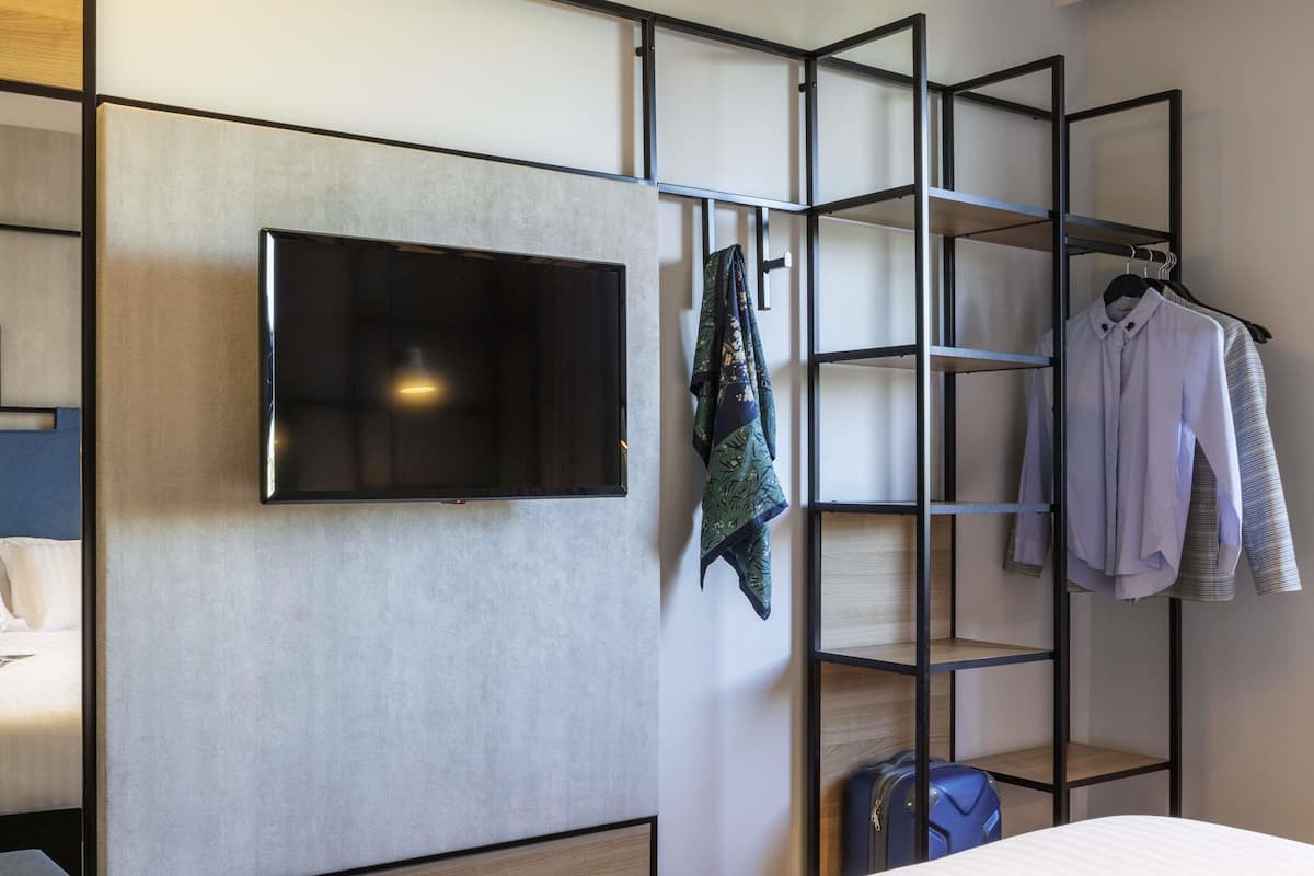 Une chambre d’hôtel équipée d’un lit, d’un téléviseur fixé au mur, d’un portant à vêtements et d’un espace de rangement pour les bagages.