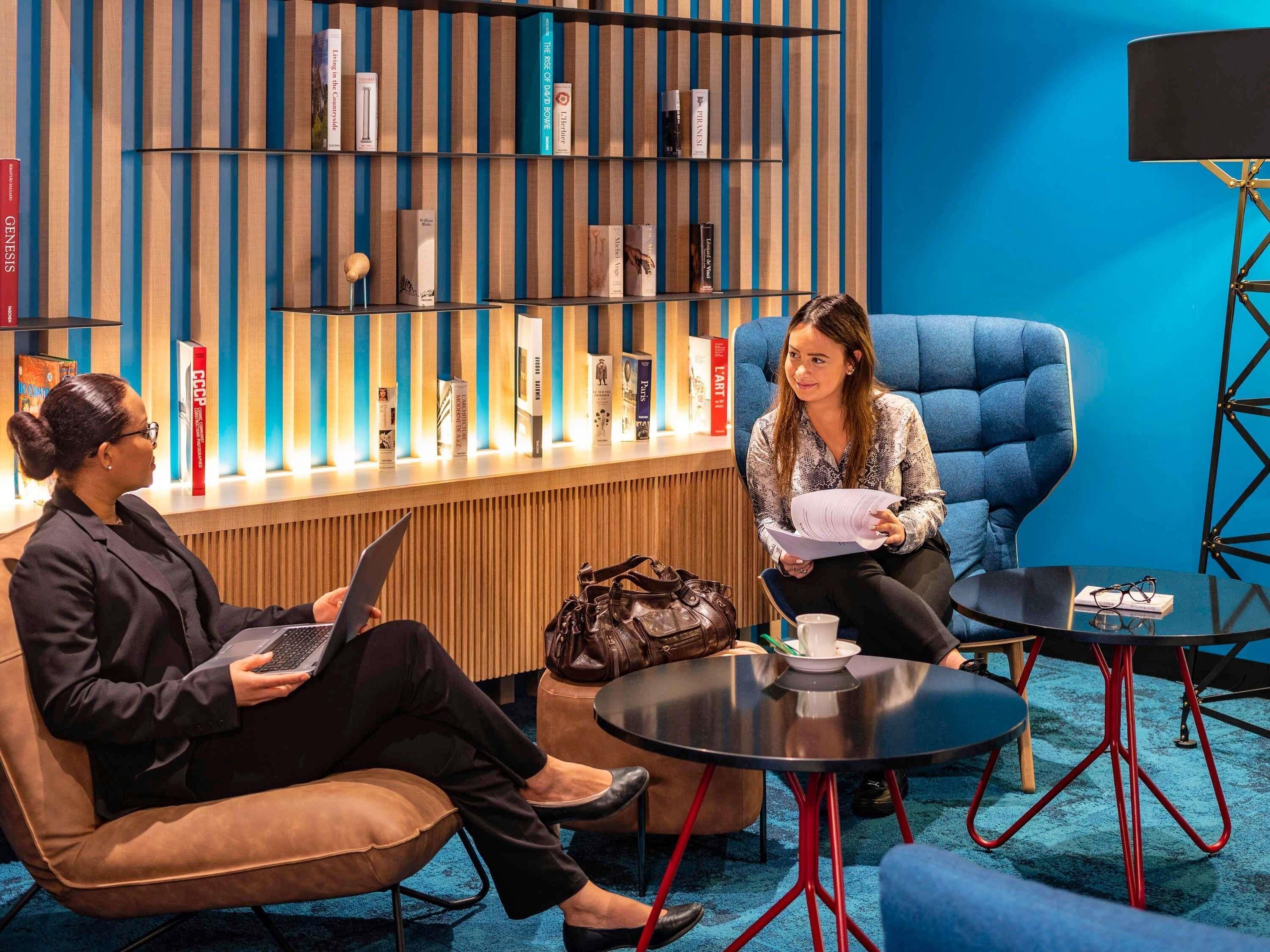 A modern lounge area with a blue wall, a bookshelf, and two seating areas with tables.