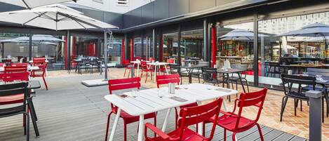 Un espacio de comedor al aire libre con mesas y sillas, grandes sombrillas y vistas de la ciudad.