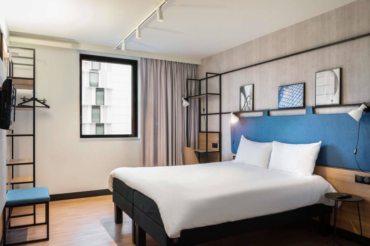 A modern hotel room with a large bed, a desk, a chair, a wardrobe, and a window with curtains.