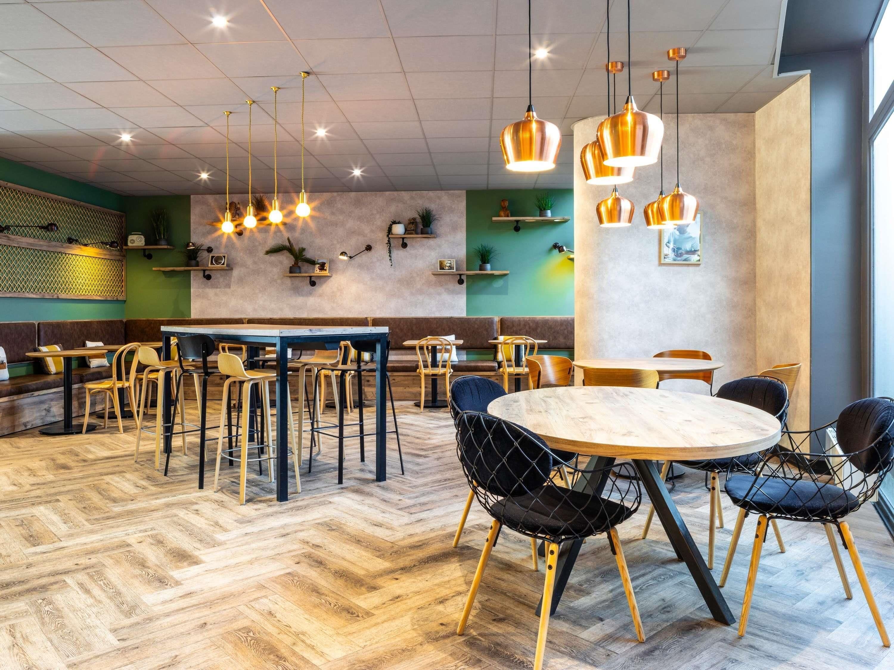 Interior de restaurante moderno com piso em padrão herringbone, mistura de mobiliário de madeira e metal, iluminação pendente e parede verde como detalhe.
