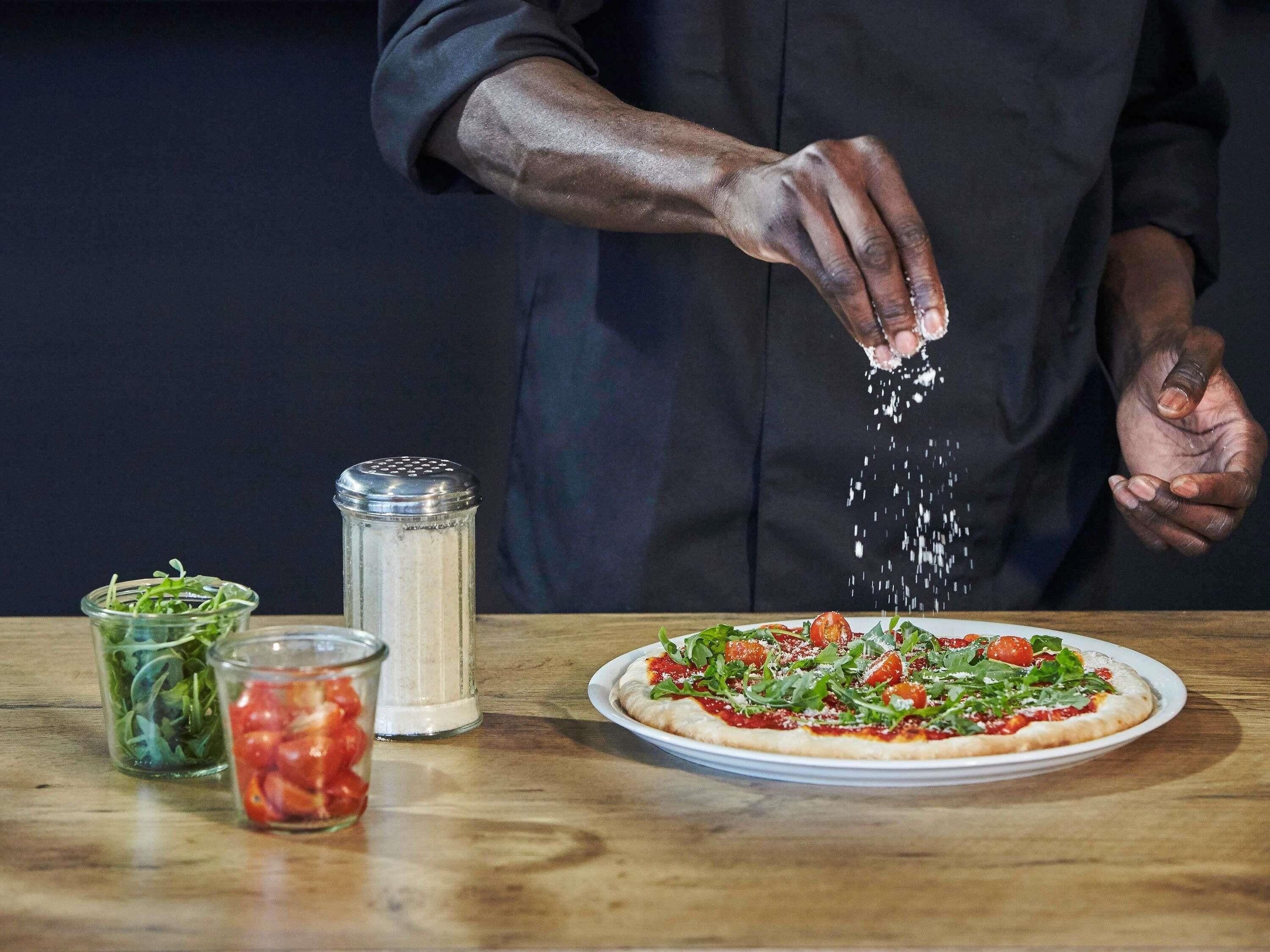 A person seasoning a pizza with various toppings and condiments on a wooden table.