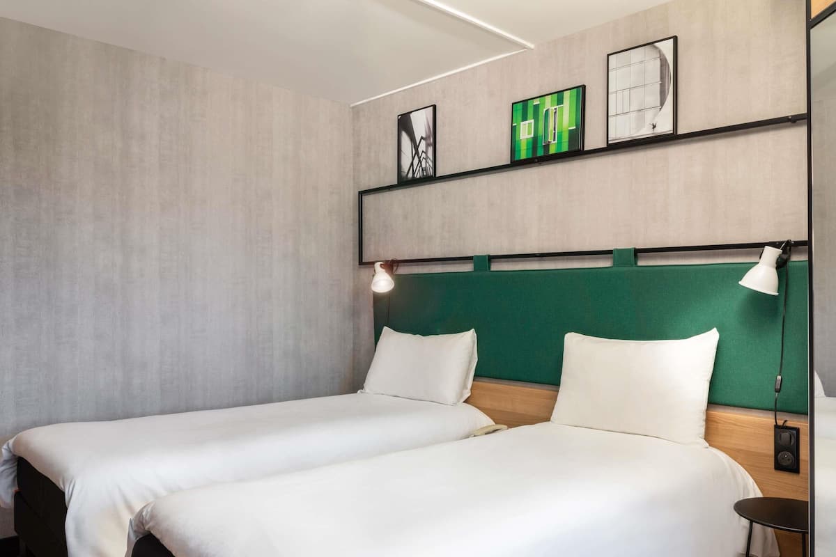 A hotel room with two beds, a green headboard, a small table, and a wall-mounted shelf with framed pictures.