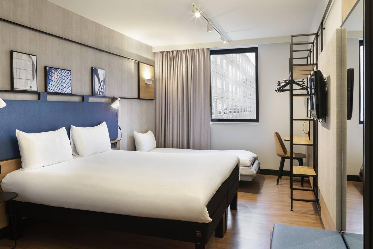 Une chambre d’hôtel avec un grand lit, un bureau, une chaise, une télévision et une fenêtre donnant sur des immeubles.