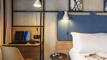 A hotel room with a bed, a desk with a laptop, headphones, and a small table with glasses and a magazine.