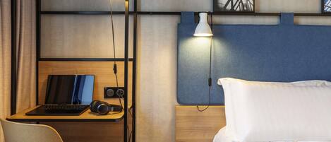 Una habitación de hotel con una cama, un escritorio con un ordenador portátil, auriculares y una mesita con vasos y una revista.