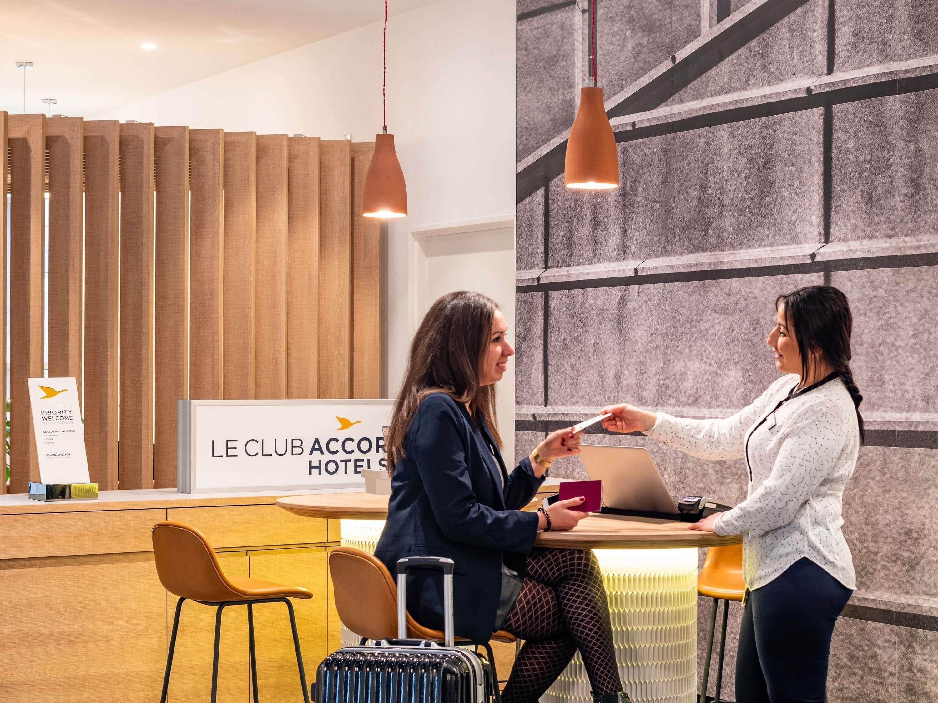 Een receptiebalie met een bord waarop 'LE CLUB ACCOR HOTELS' staat, een houten lambrisering en een moderne hanglamp.