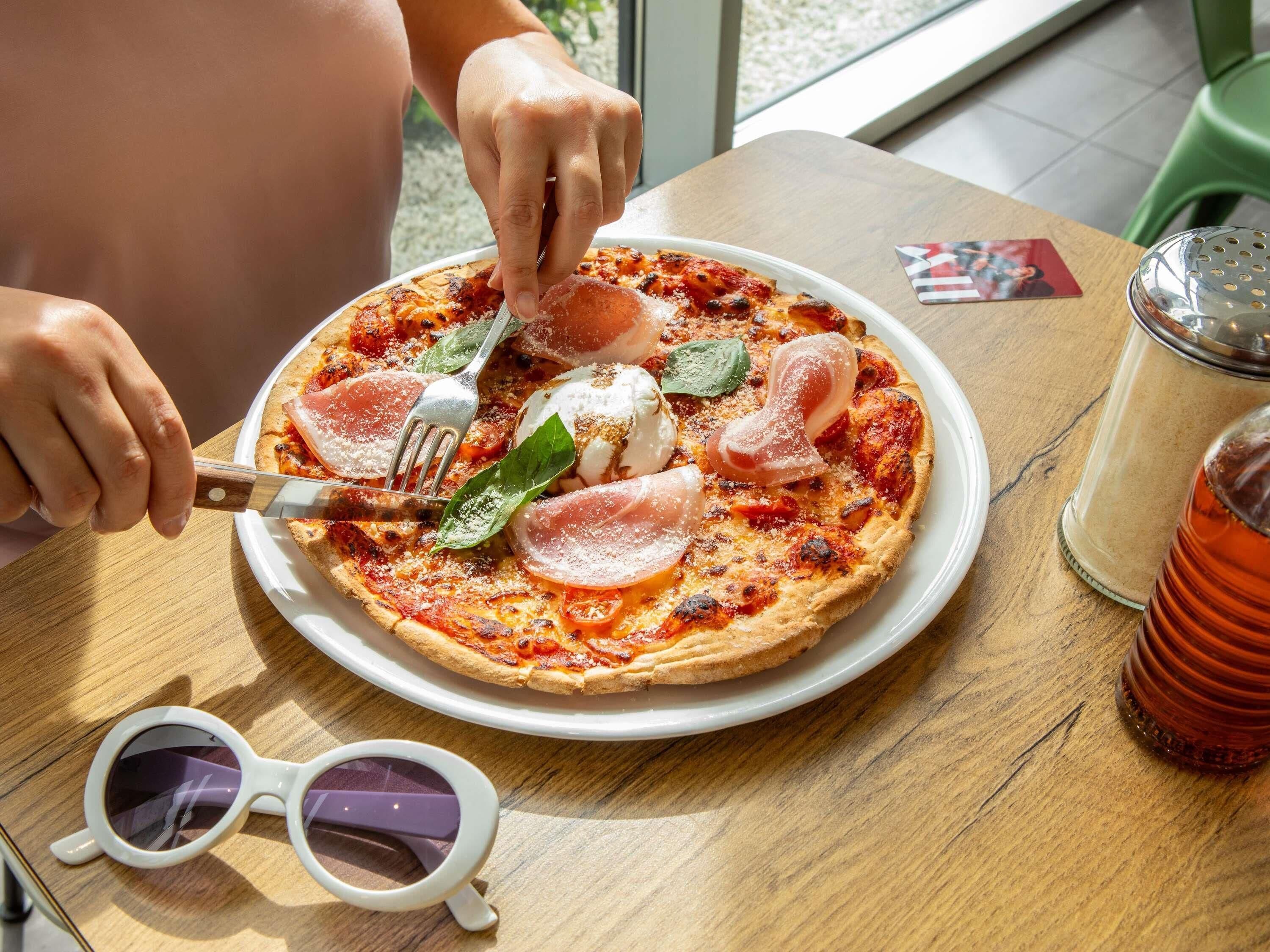 A pizza with prosciutto and mozzarella on a white plate, a pair of sunglasses, and a glass of red drink on a wooden table.