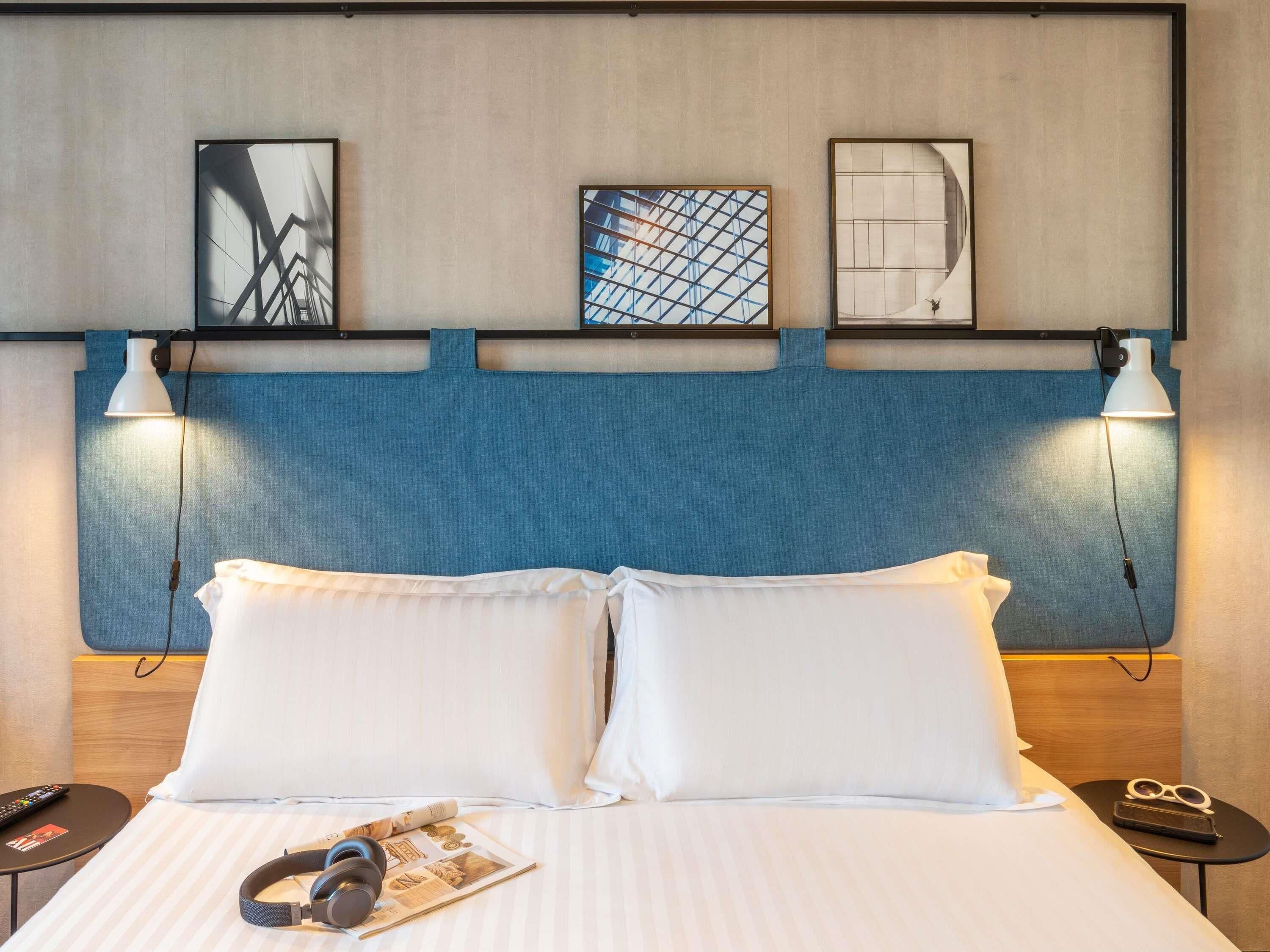 Ein Hotelzimmer mit einem Bett, zwei Nachttischlampen, einer Kopfteil und drei gerahmten Kunstwerken an der Wand.