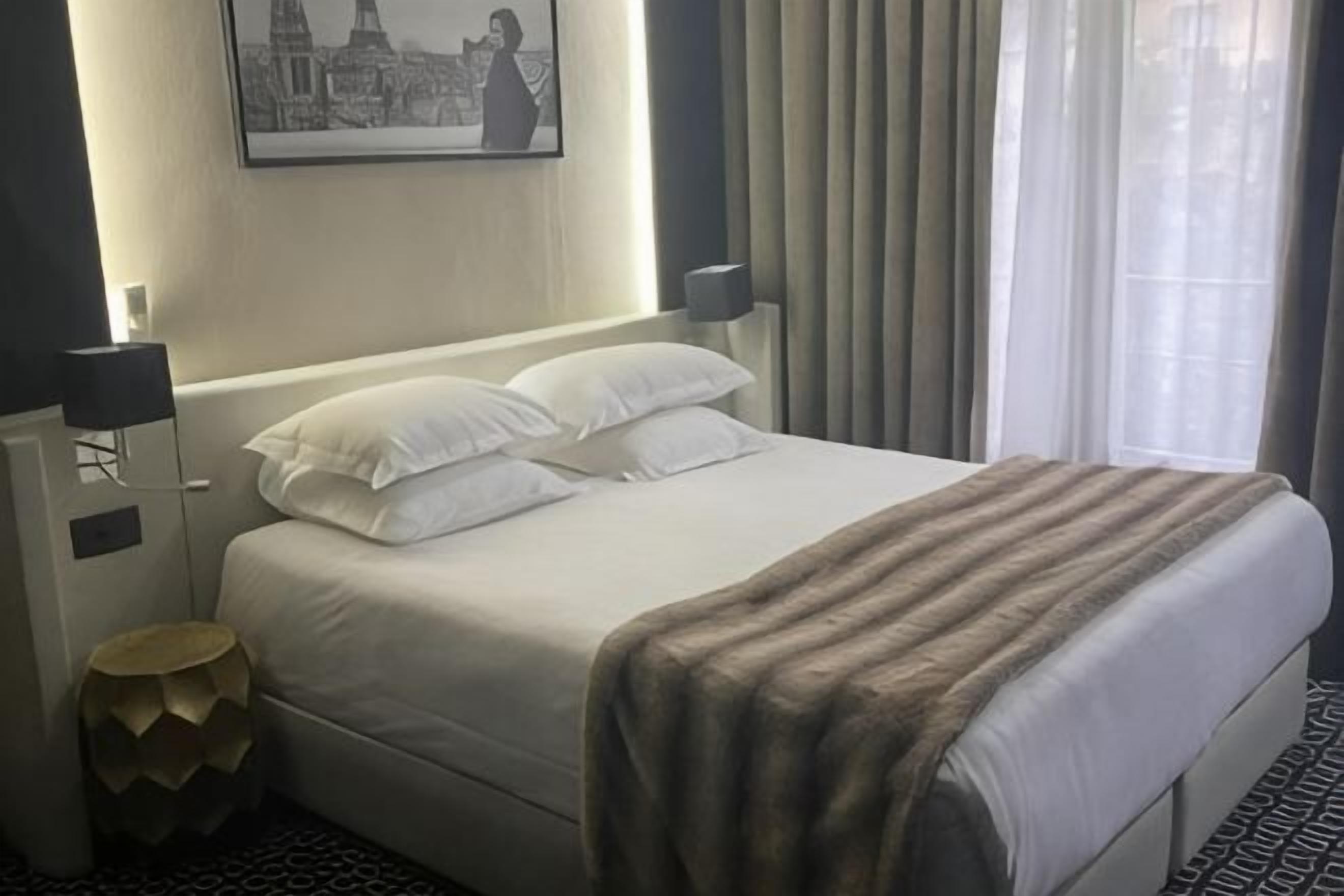 A hotel room with a bed, a bedside lamp, a wall-mounted picture, and a window with curtains.