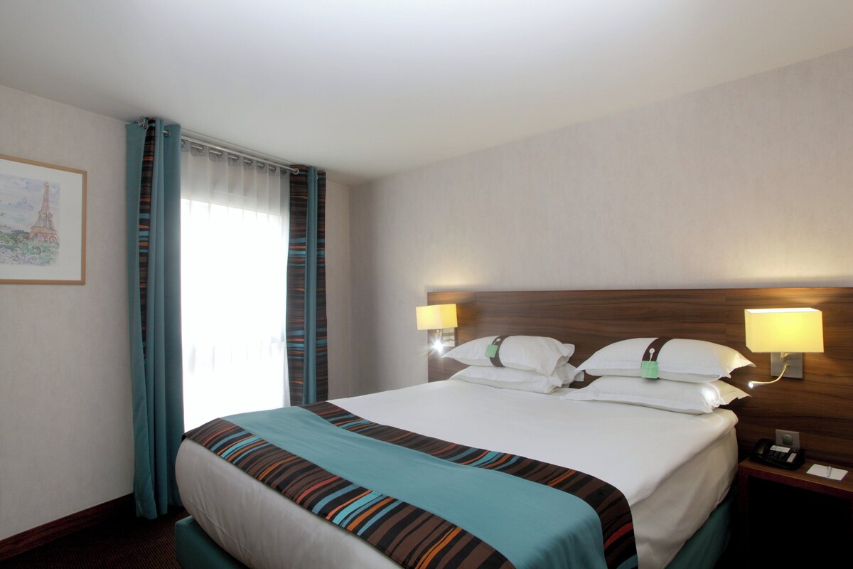Quarto de hotel com cama, duas luminárias de cabeceira, mesa de cabeceira, telefone e janela com cortinas.
