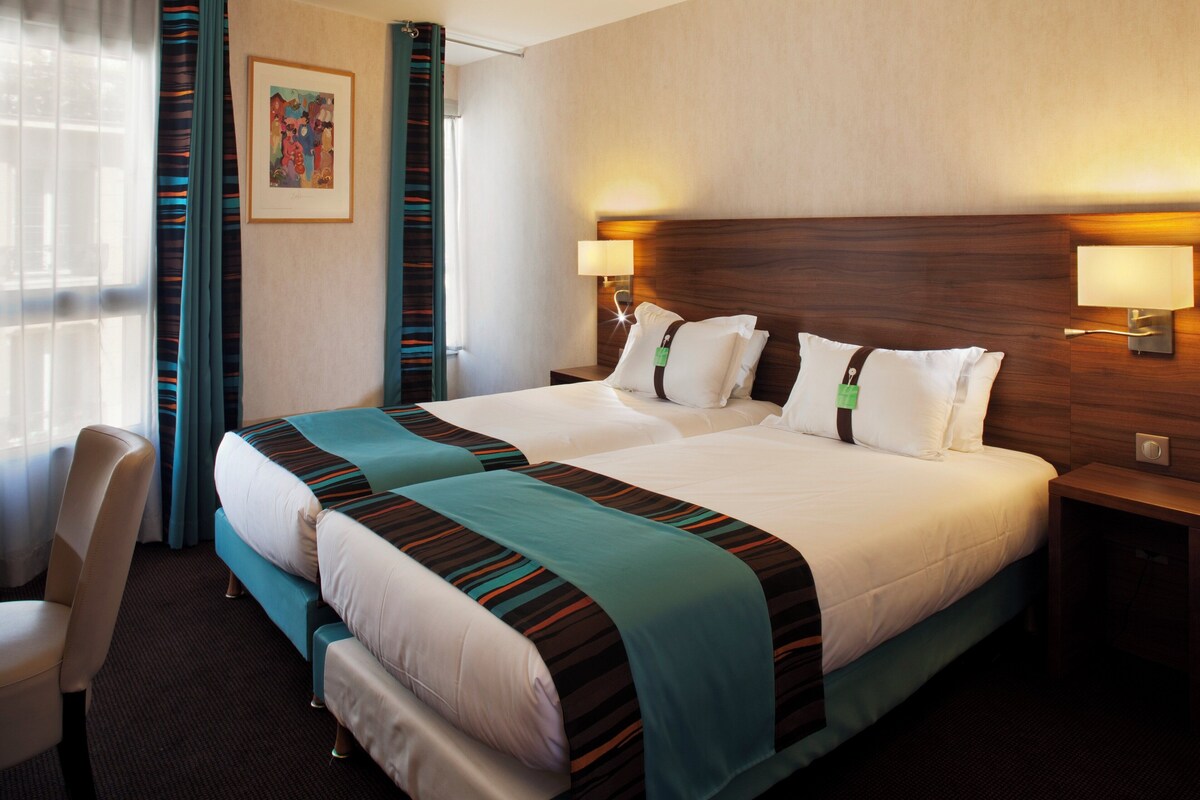 Quarto de hotel com uma cama grande, duas mesas de cabeceira, uma escrivaninha e uma cadeira.