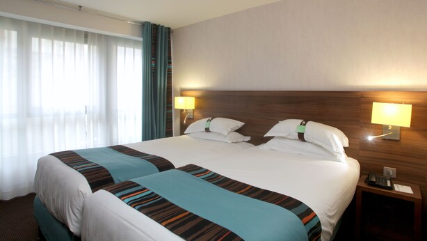 A hotel room with a large bed, two bedside tables, a nightstand, a lamp, and a window with curtains. - Holiday Inn Paris - Montmartre by IHG (Paris)