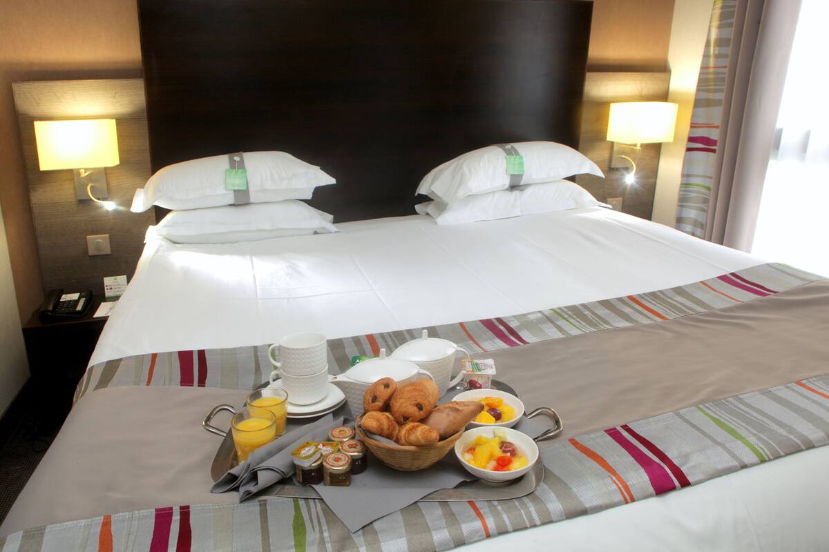 A hotel room with a neatly made bed, breakfast tray with croissants, fruit, and juice, and a bedside table with a lamp and a phone.