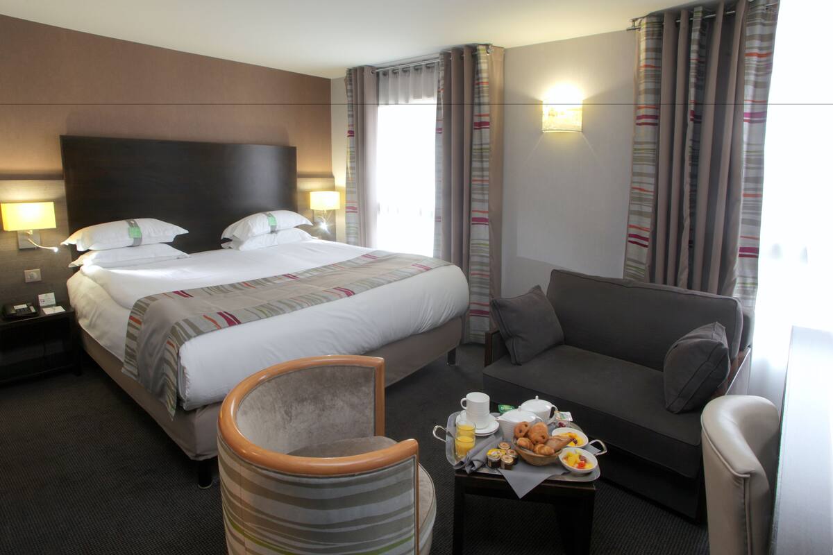 A hotel room with a large bed, a sofa, a chair, and a breakfast tray with pastries and coffee.