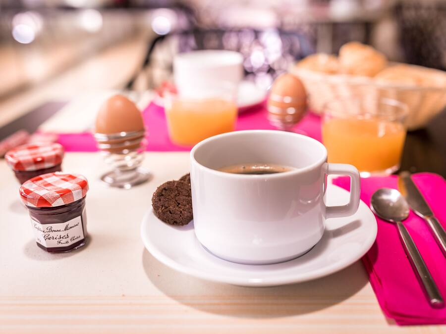 Ein Frühstückstisch mit einer Tasse Kaffee, einem Glas Orangensaft, Eiern, einem Marmeladenglas und einem Korb Croissants auf einer rosa Servietten.