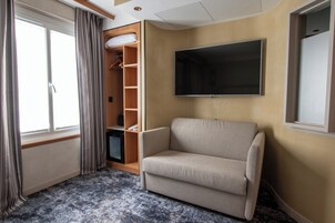 A room with a sofa, a TV mounted on the wall, a window with curtains, and a wooden wardrobe.