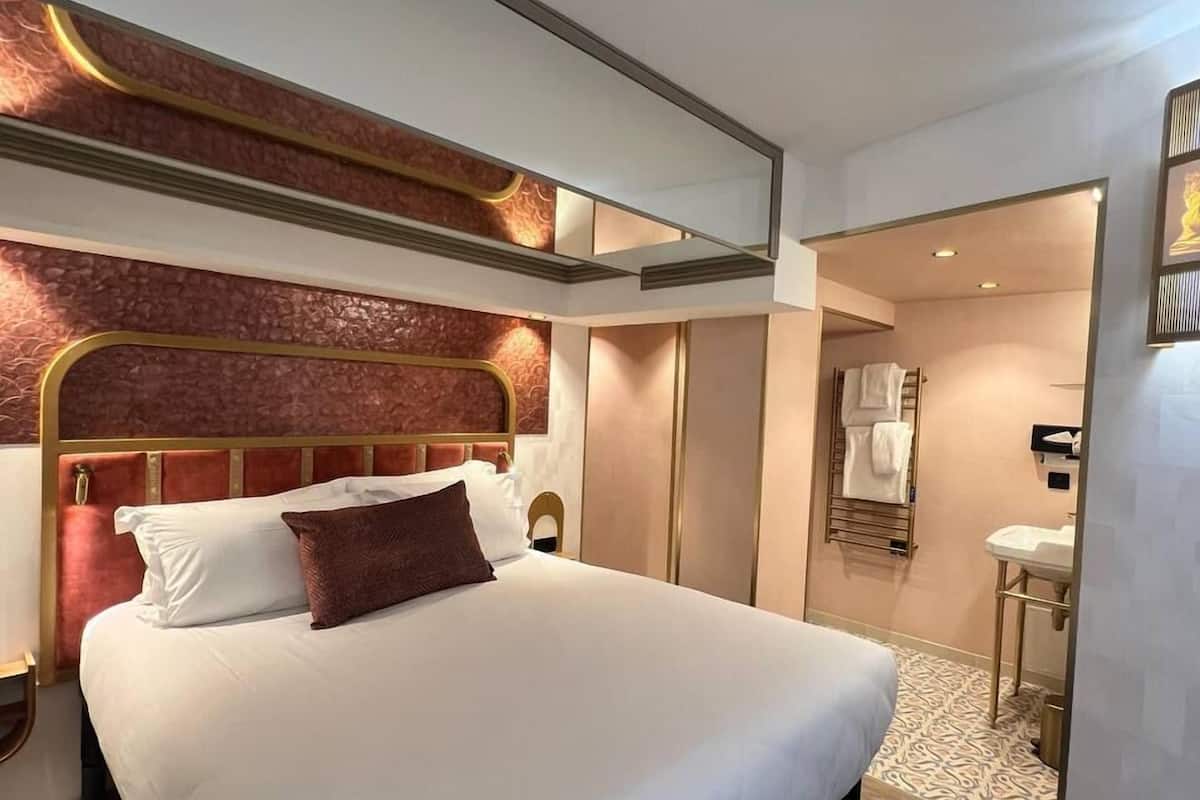 Una habitación de hotel moderna con una cama grande, un cabecero de diseño único, una mesita de noche y un baño visible a través de una puerta abierta.