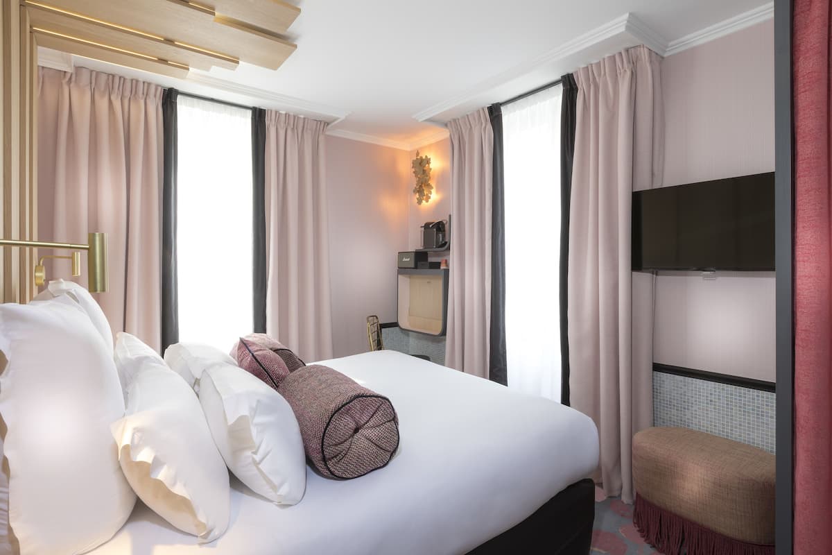 Hotellihuone, jossa on suuri sänky, seinälle asennettu taulutelevisio, pieni oleskelualue ja ikkuna, jossa on verhot.
