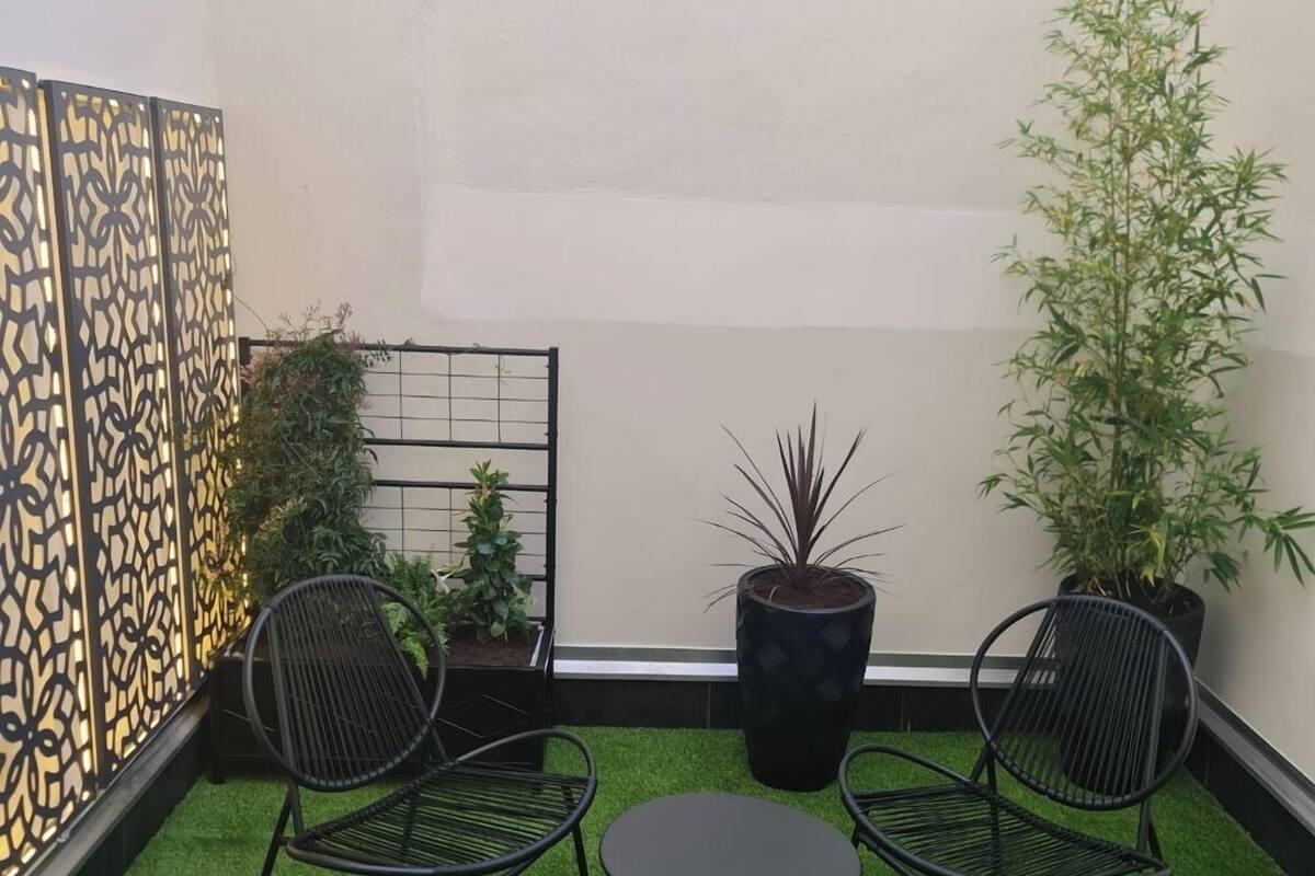 Un pequeño patio con césped artificial verde, dos sillas de metal negras, una mesa redonda y plantas en macetas.