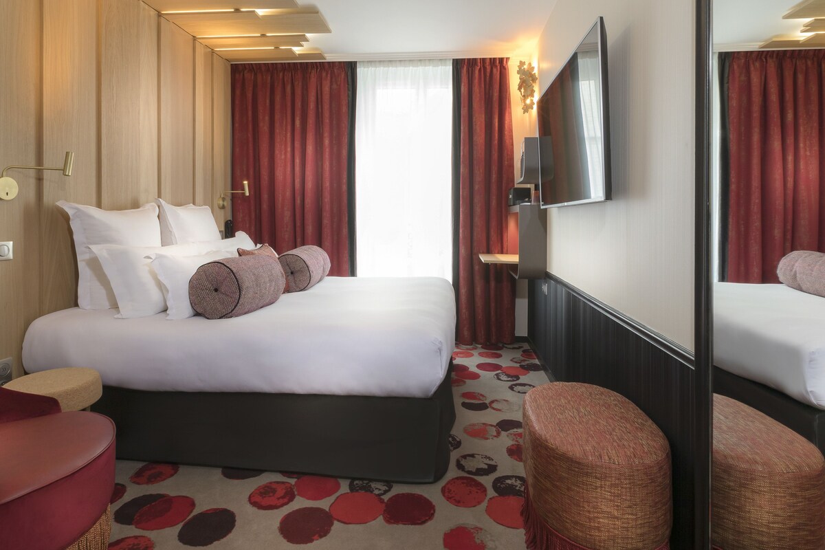 Habitación de hotel con una cama grande, televisión de pantalla plana, un espejo y una alfombra roja con diseños circulares.