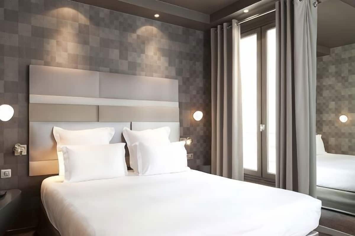 Una habitación de hotel moderna con una cama grande, un cabecero con iluminación integrada, un ventanal con cortinas y un espejo que refleja la habitación.