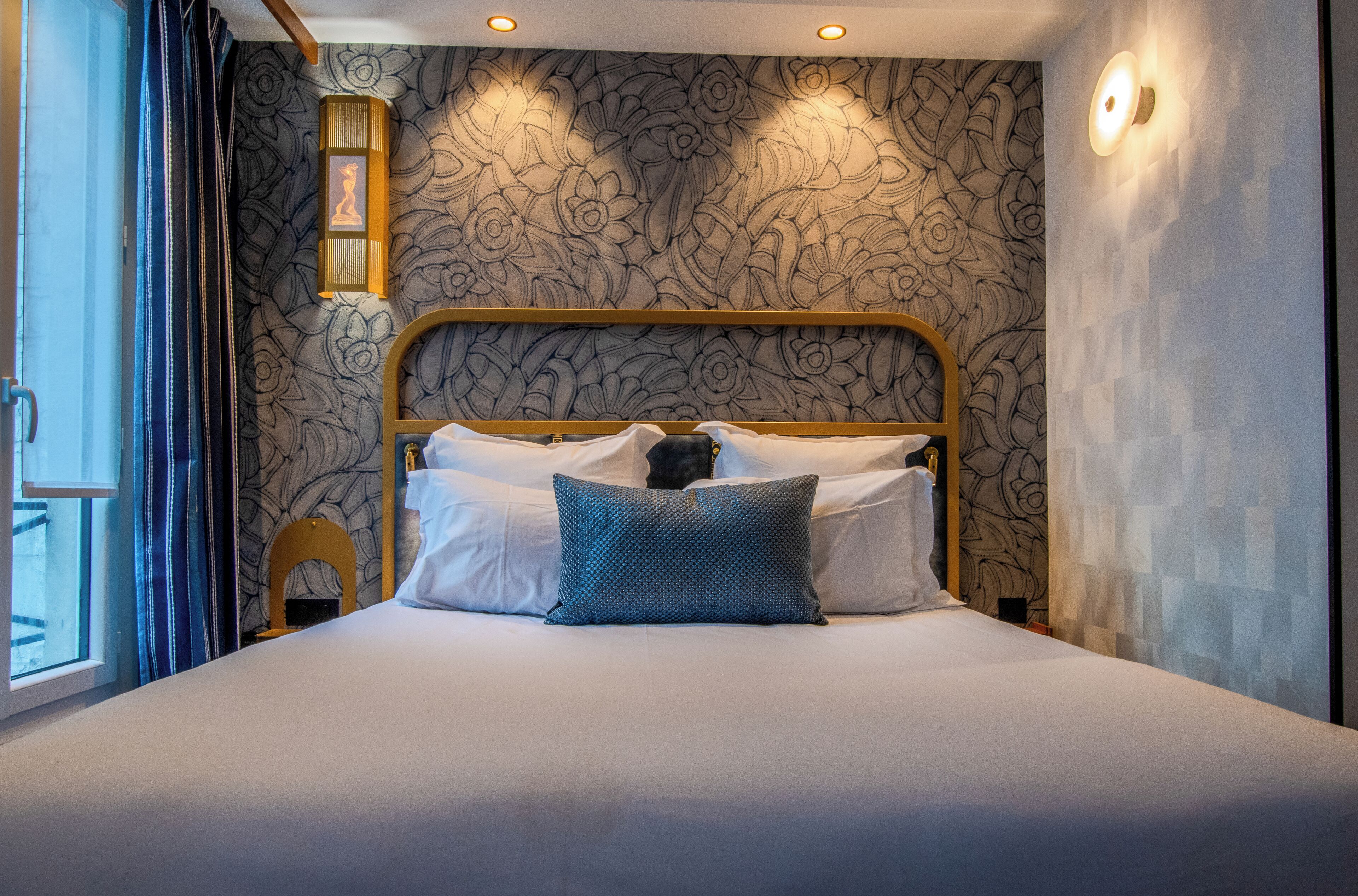 Cama bem arrumada com almofadas brancas e azuis, cabeceira de madeira e papel de parede com padrão floral.