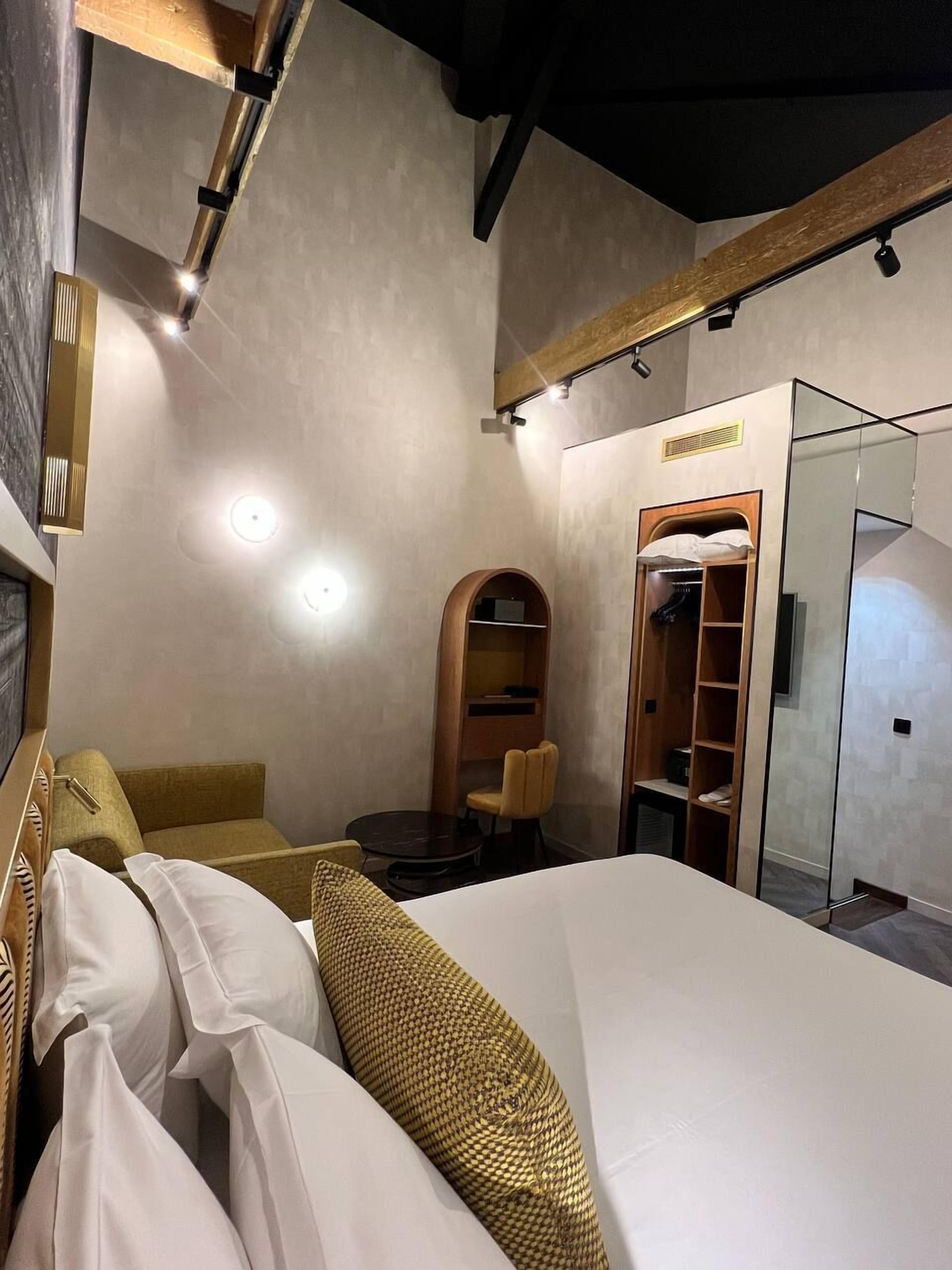 A hotel room with a large bed, a wooden headboard, a small seating area with a chair and table, a wardrobe, and a mirror.