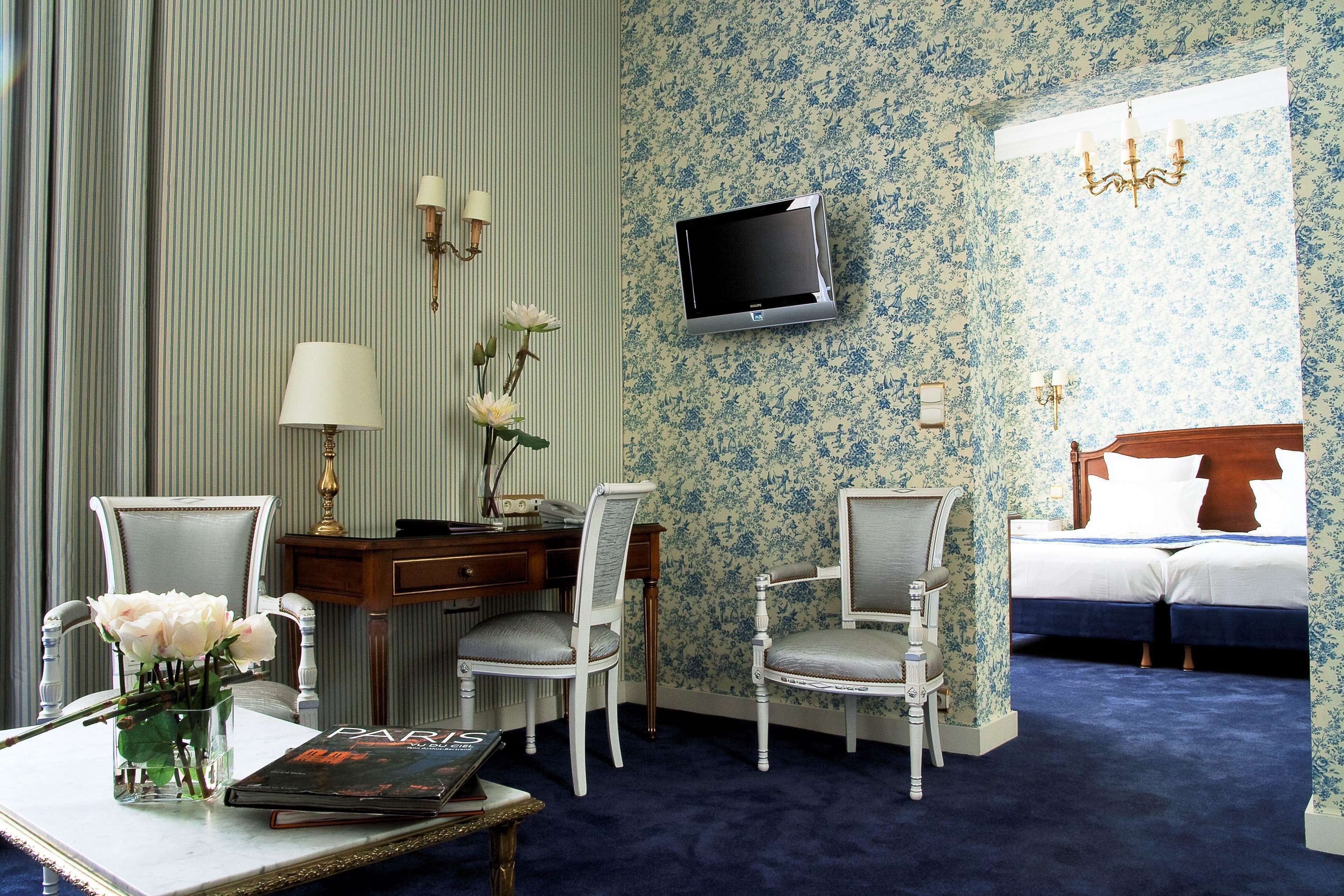 Hotelkamer met een bed, twee stoelen, een bureau, een televisie en een vaas met bloemen.