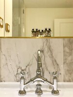 A bathroom with a marble sink, a mirror above it, and a shelf with bottles.