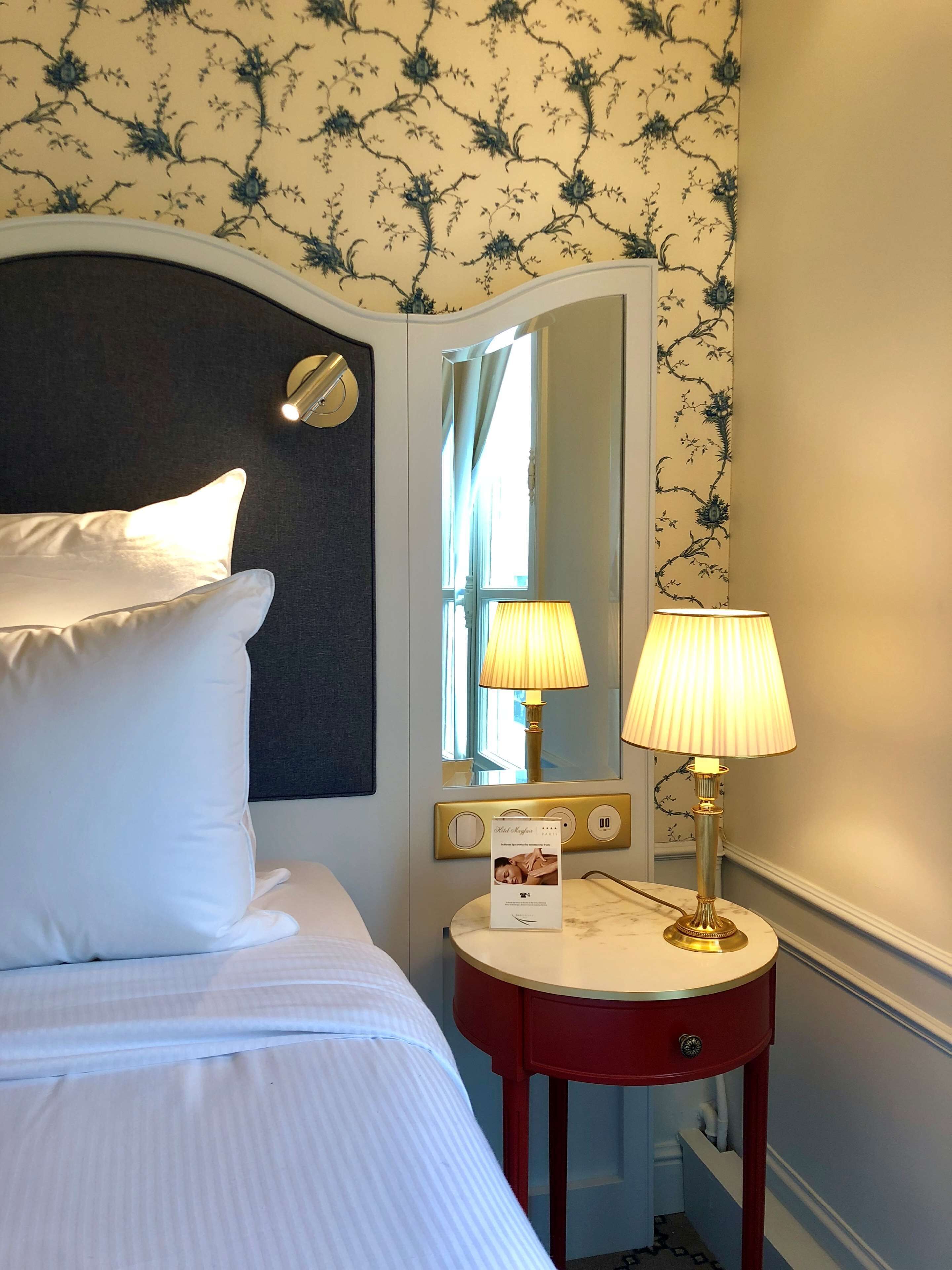 A hotel room with a bed, a bedside table with a lamp, a mirror, and floral wallpaper.