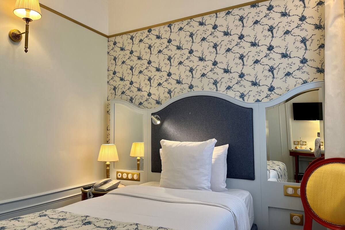 A hotel room with a bed, bedside lamps, a mirror, a television, and a chair.