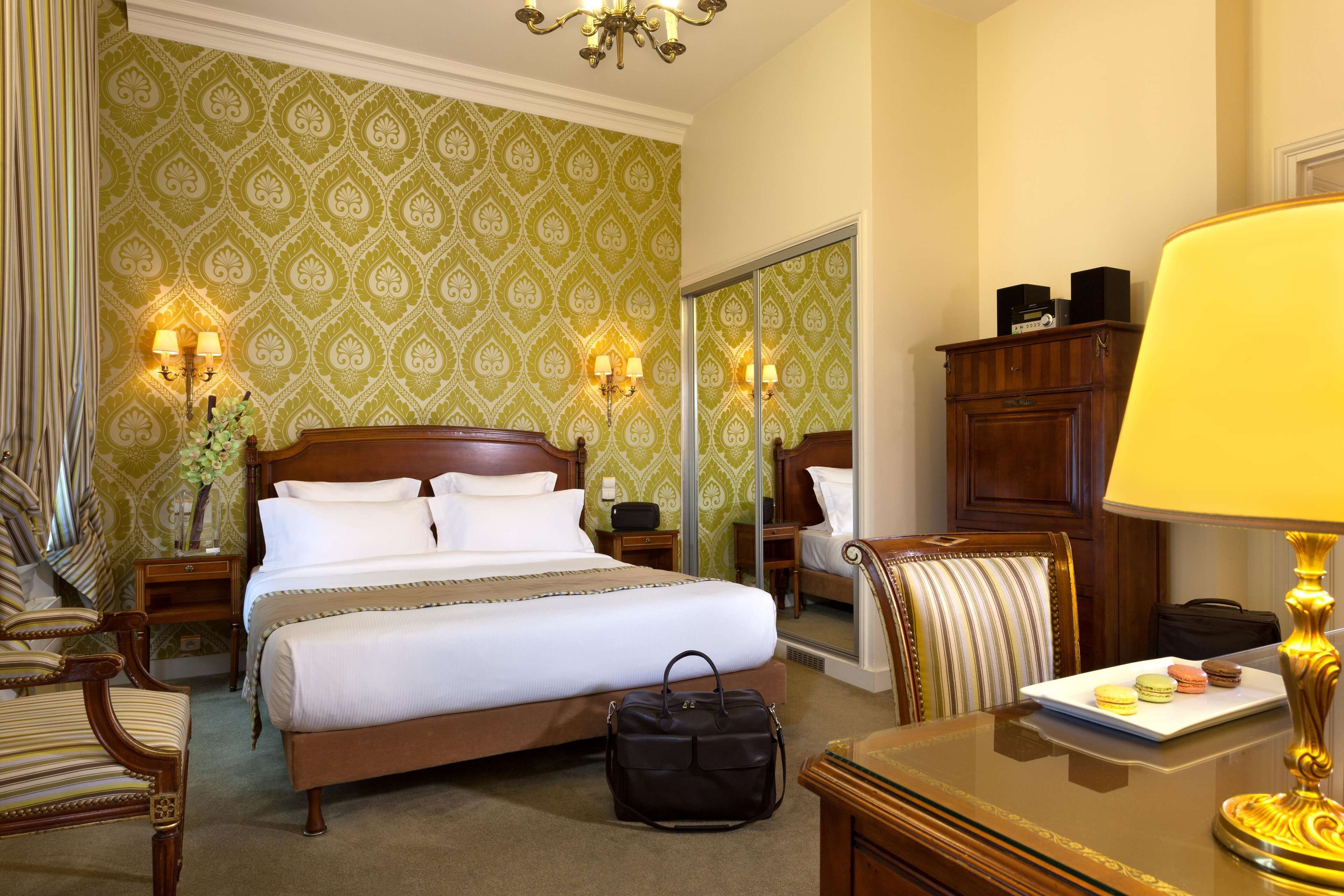 A hotel room with a large bed, bedside tables, a chair, a desk with a lamp, and a mirror.