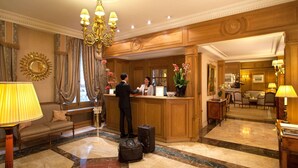 A lobby with a reception desk, a sofa, a chandelier, and a large mirror. - Hotel Mayfair (Paris)