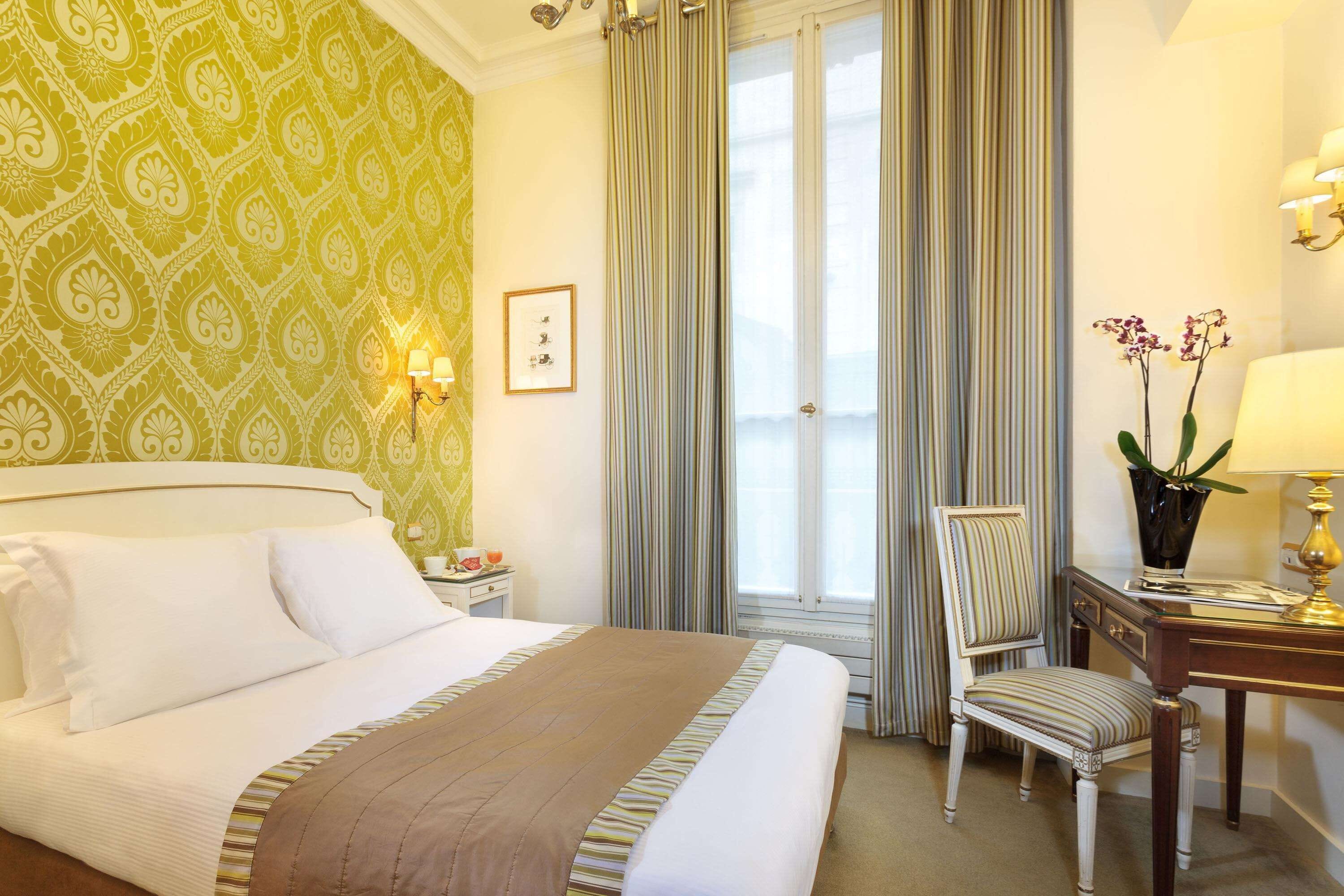 A hotel room with a large bed, a desk with a lamp, a chair, a potted plant, and a window with curtains.