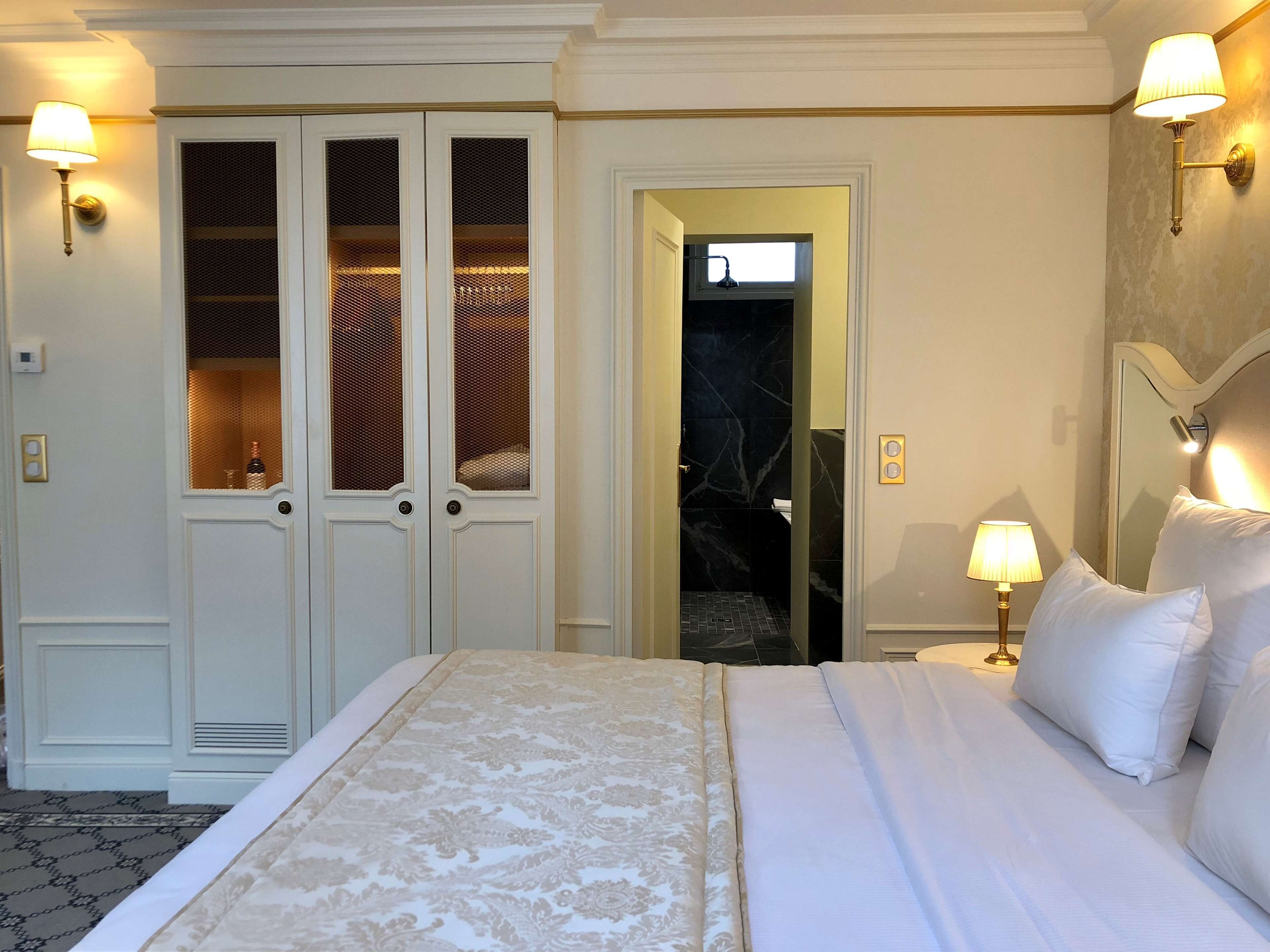 Une chambre d’hôtel avec un grand lit, une armoire aux portes vitrées, une salle de bain visible à travers une porte ouverte et des lampes fixées au mur.